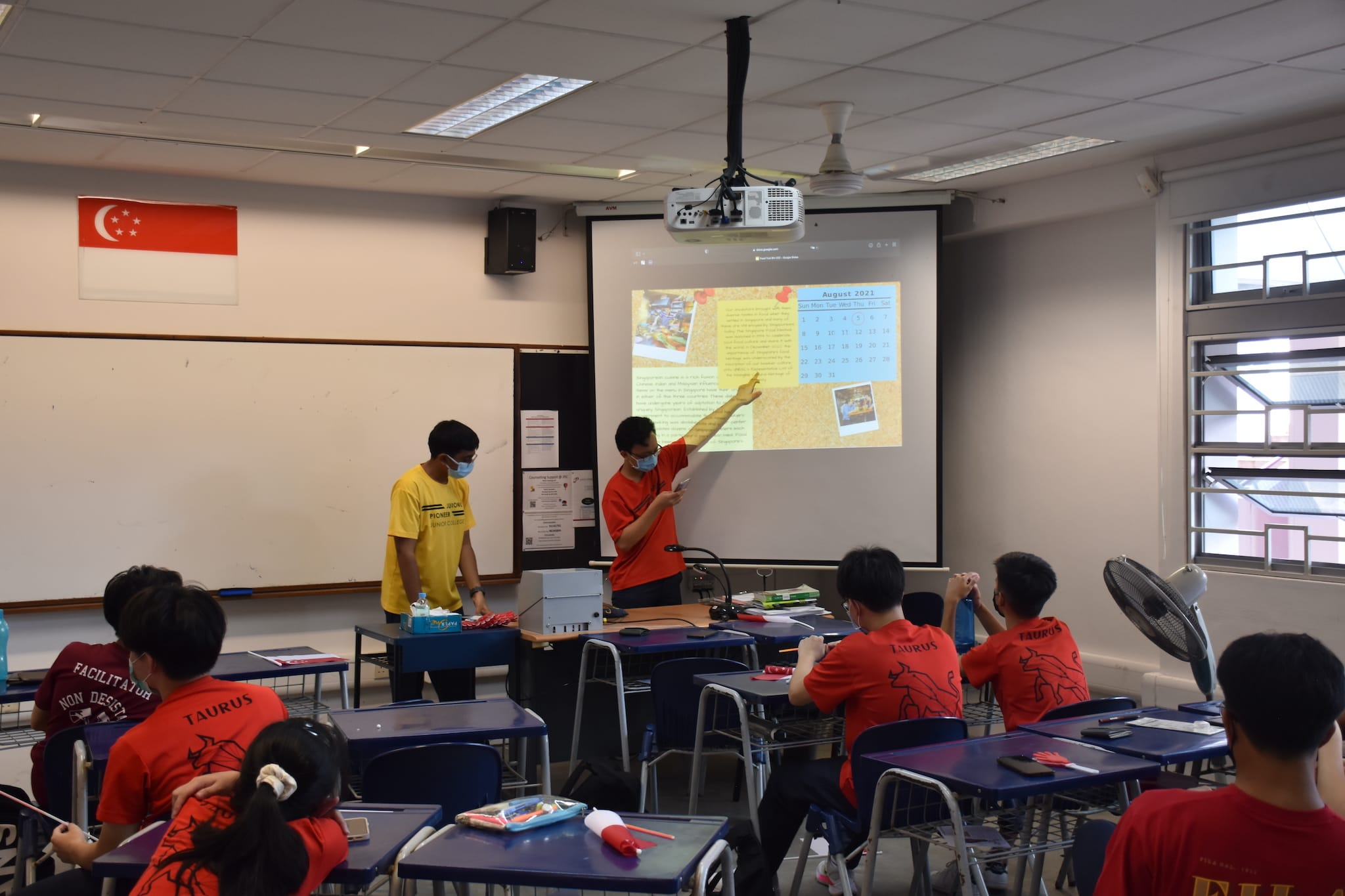 Two masked students present to classmates in a Singapore classroom, pointing at a projected August 2021 calendar slide.